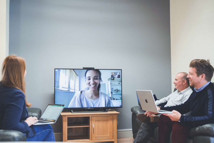 A video call between the senior management team and a remote working colleague via video call. The colleague appears on a large screen, all are smiling and have laptops.