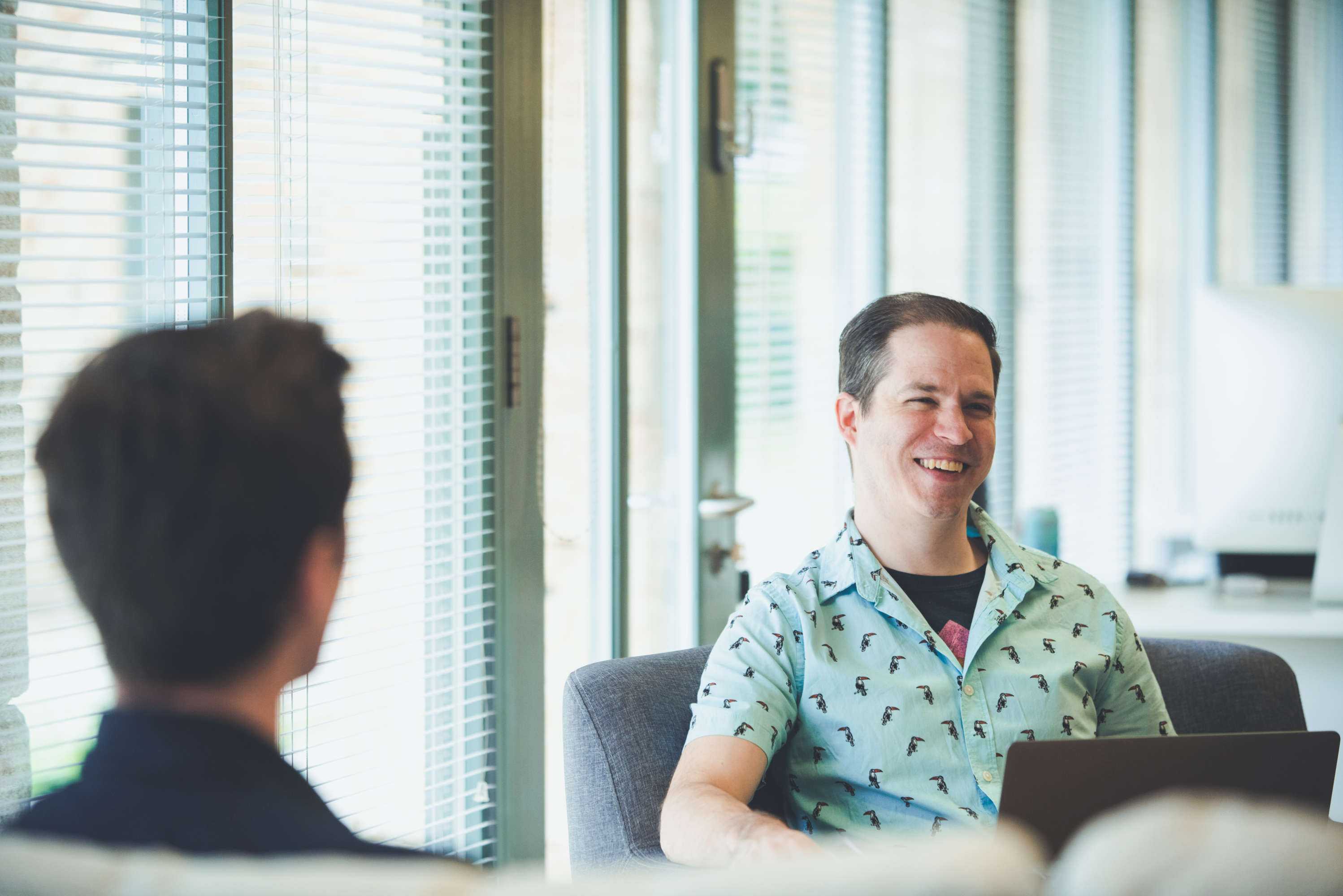 Senior designer with laptop smiles and chats with colleagues on a large sofa chair.