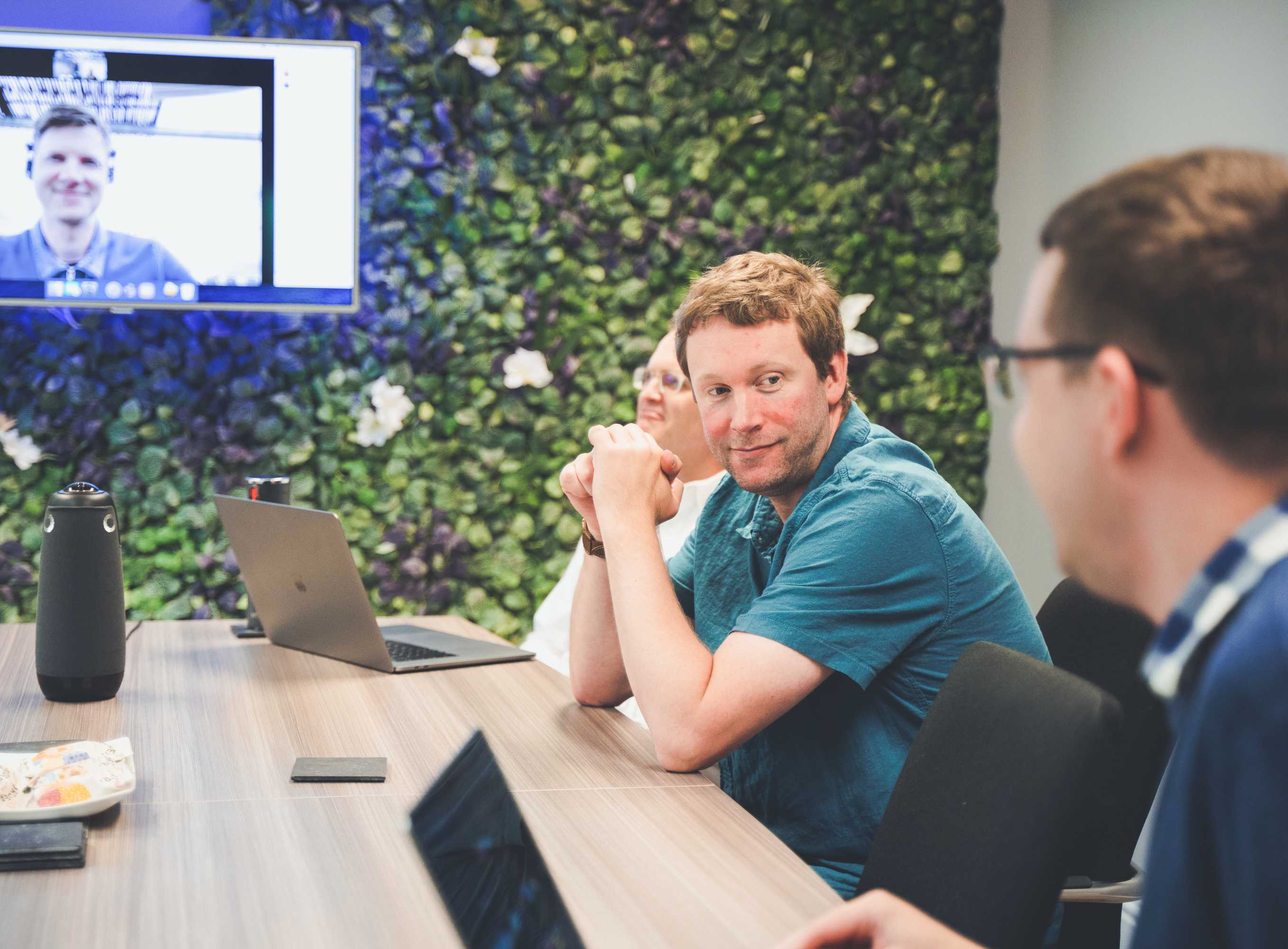 Three developers sit at a large desk with laptops, and chat with their colleague on a video call.