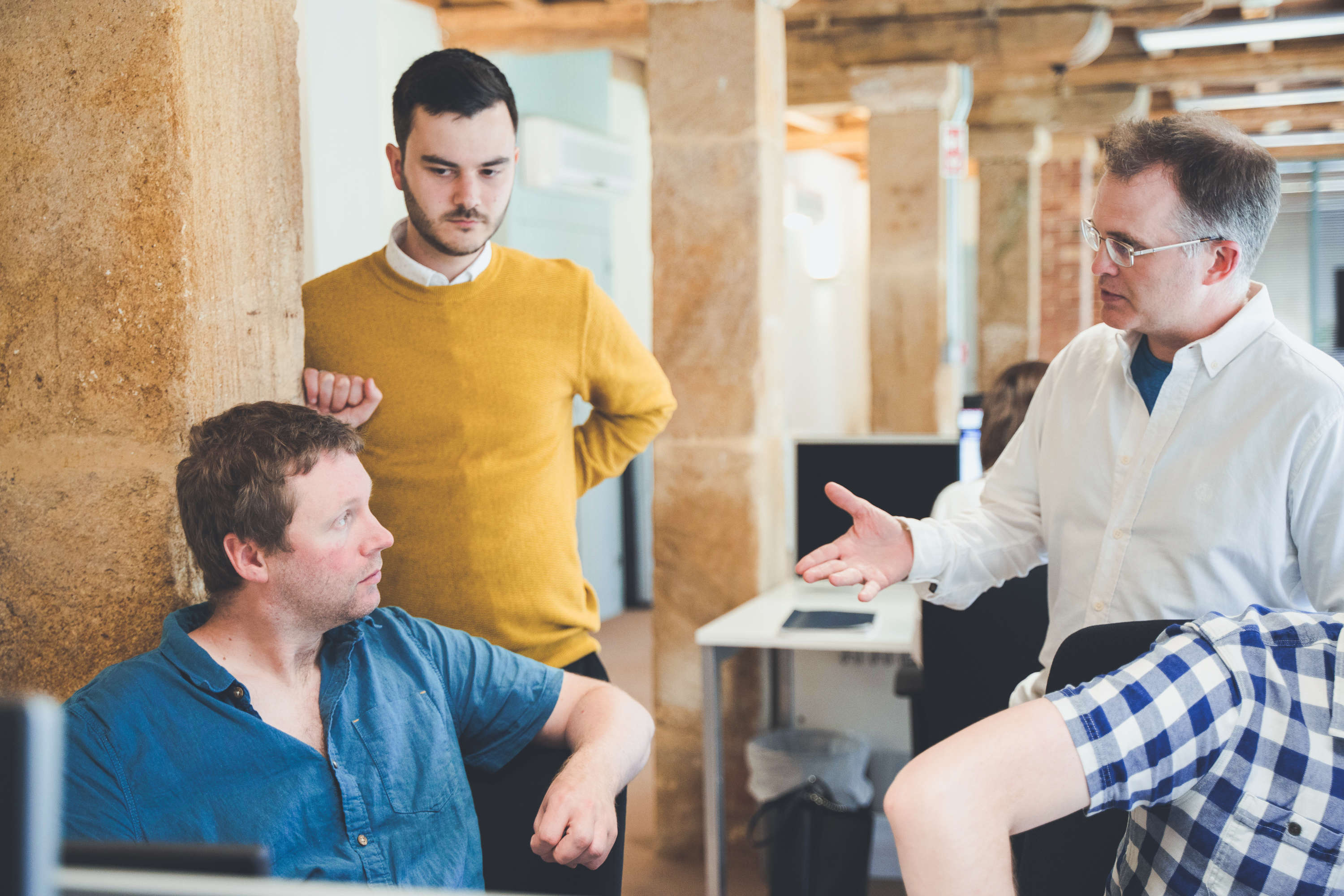Two developers and a project manager casually discuss a project, with one leaning against a stone pillar in a characterful office space.