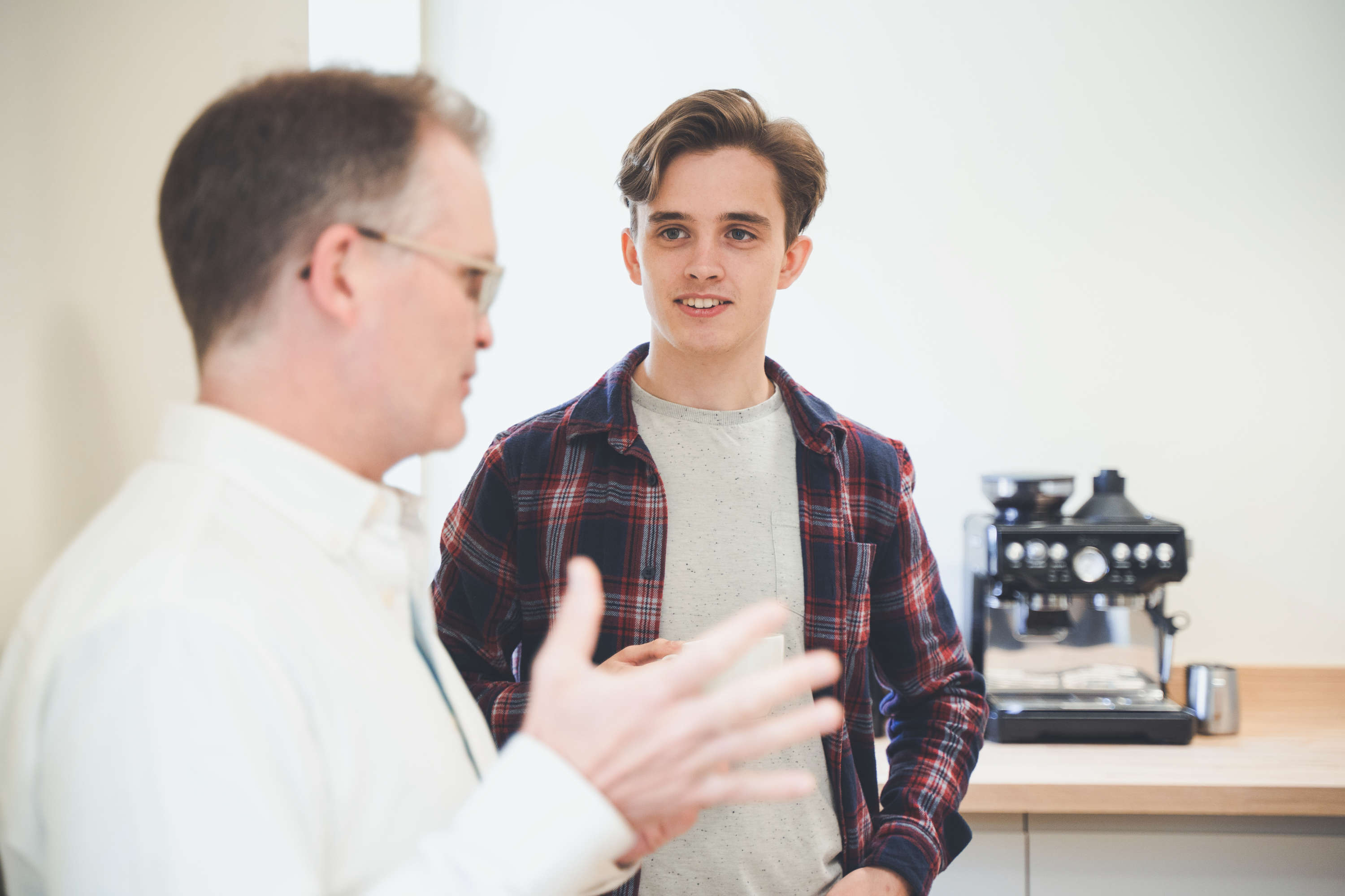 Two developers catchup during a break in the office kitchen with a coffee maker in the background.