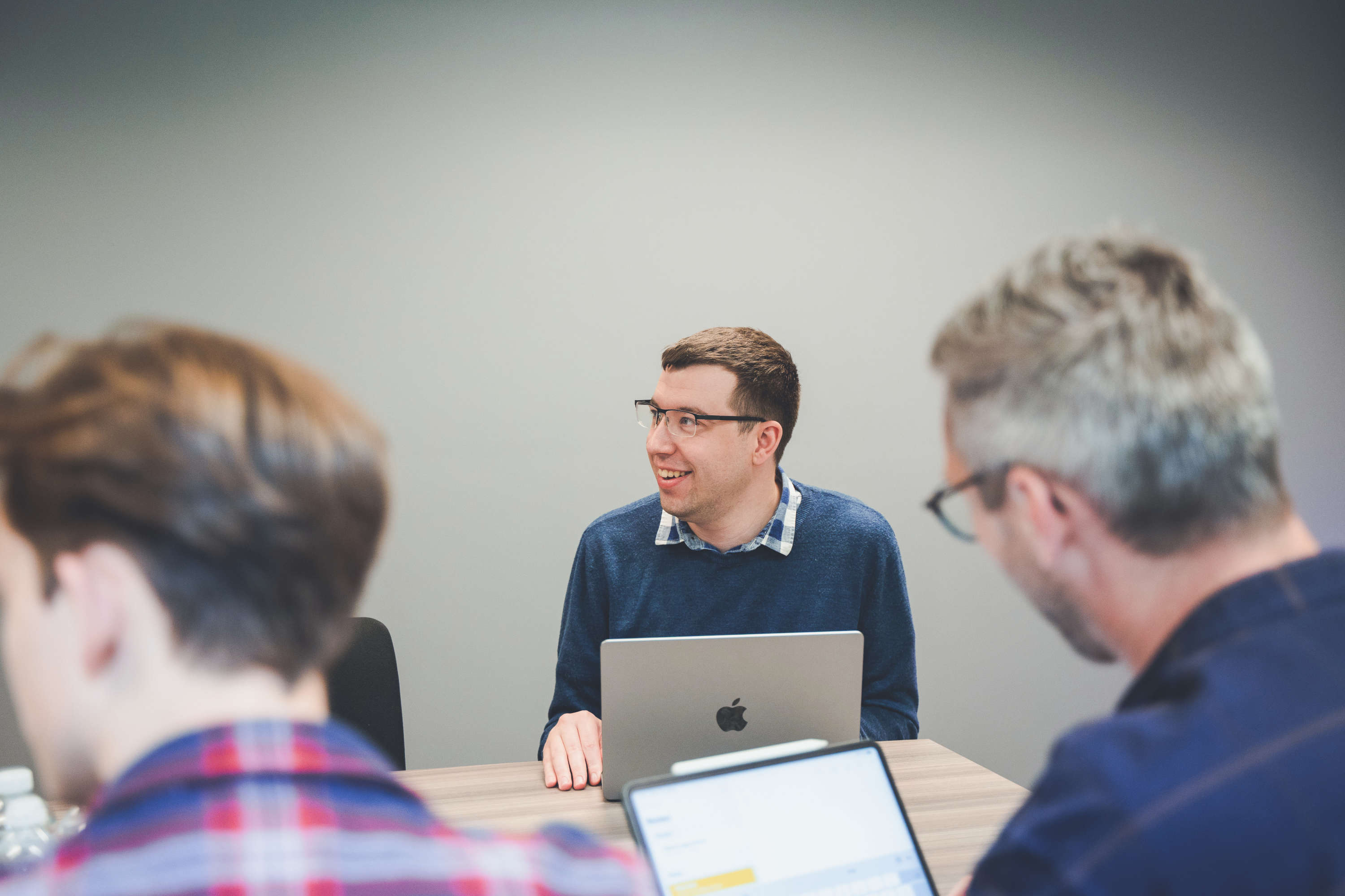 A smiling developer sits at a large desk during a team meeting, looking at a screen.