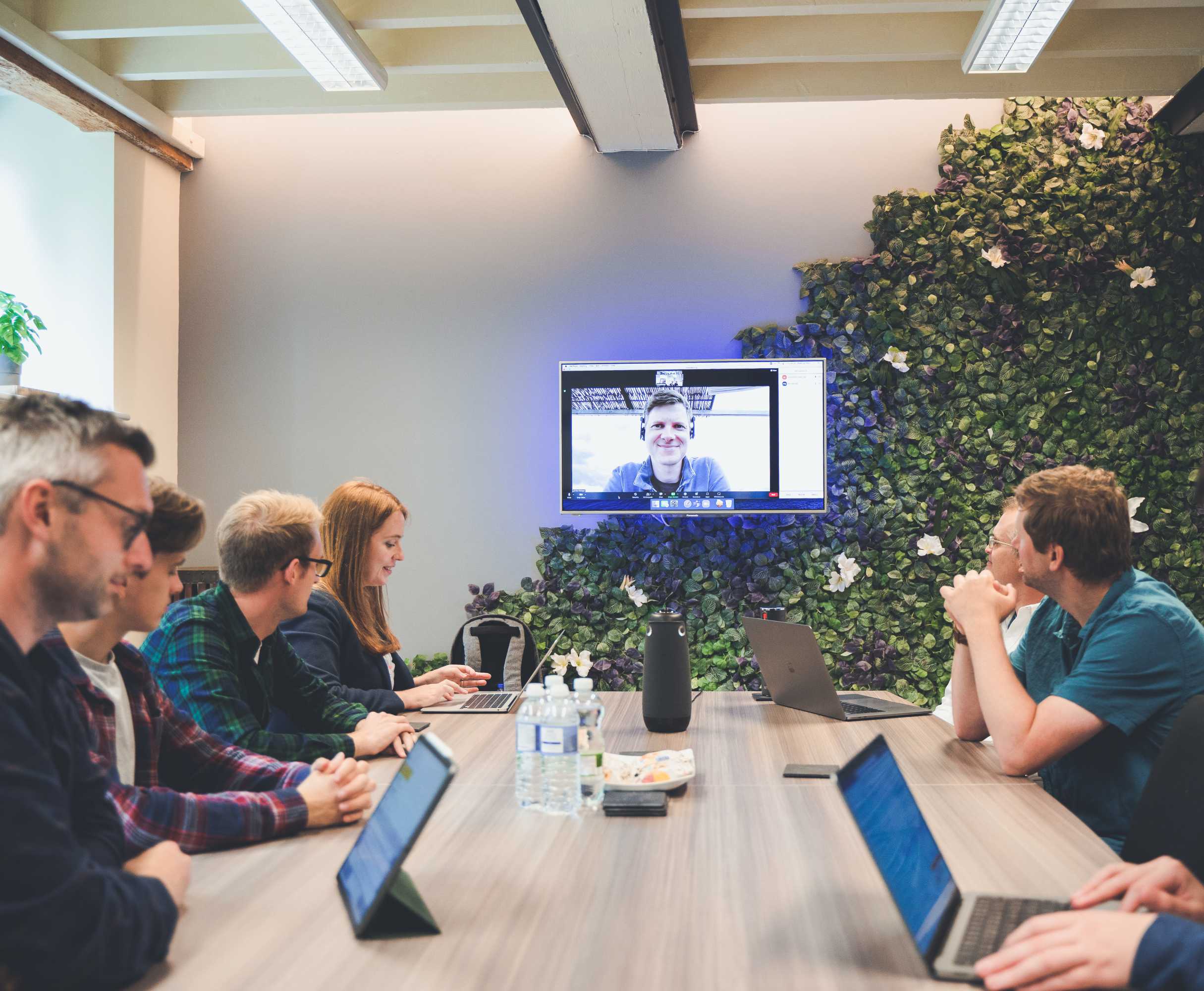 A web development team conduct a video call meeting with a colleague around a large desk. Their colleague is appearing remotely via a screen mounted on a plant-covered feature wall.