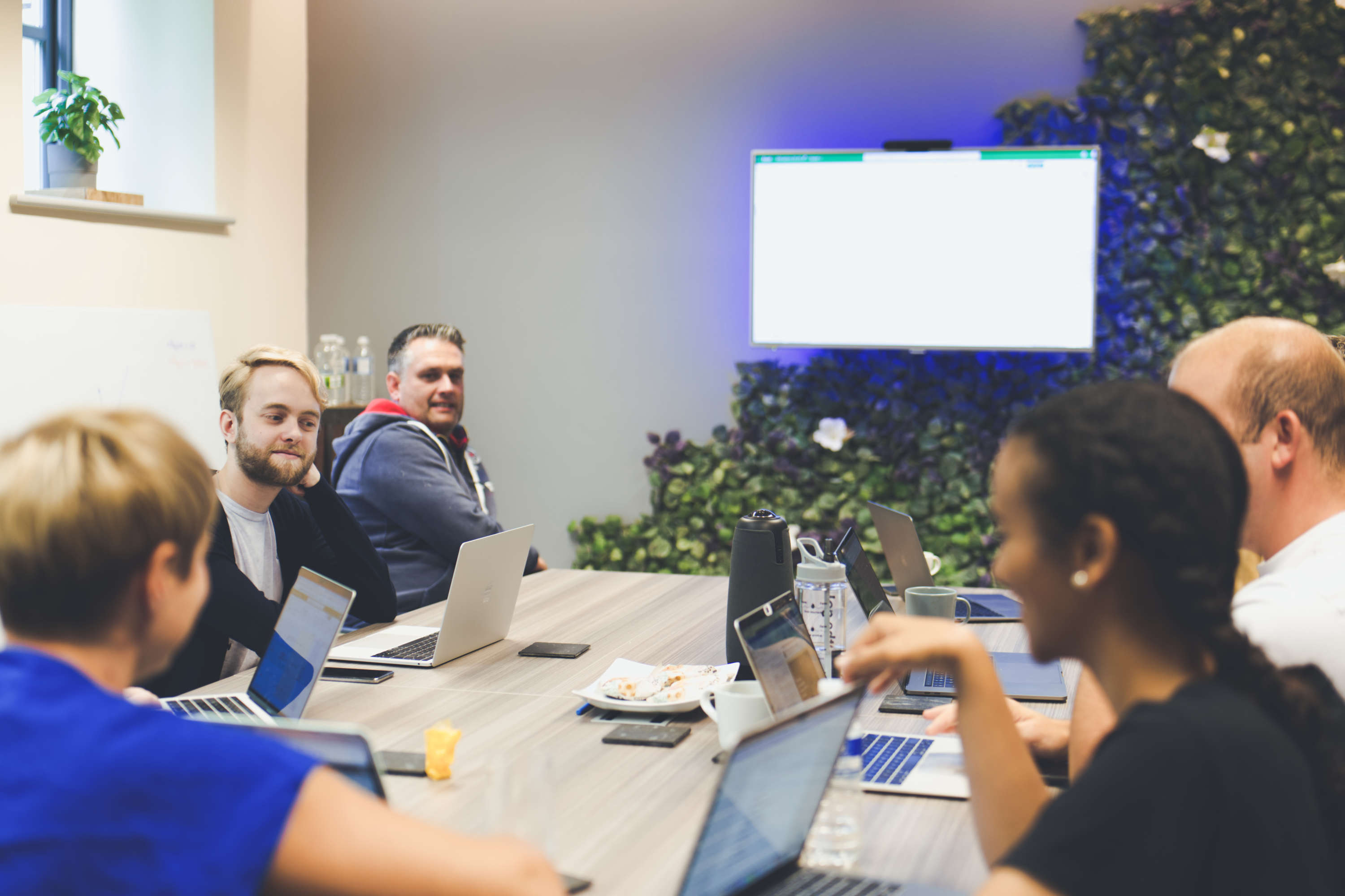 During a large team meeting, digital marketers sat around a large desk with laptops discuss their work. A large screen in the background is used to share information.