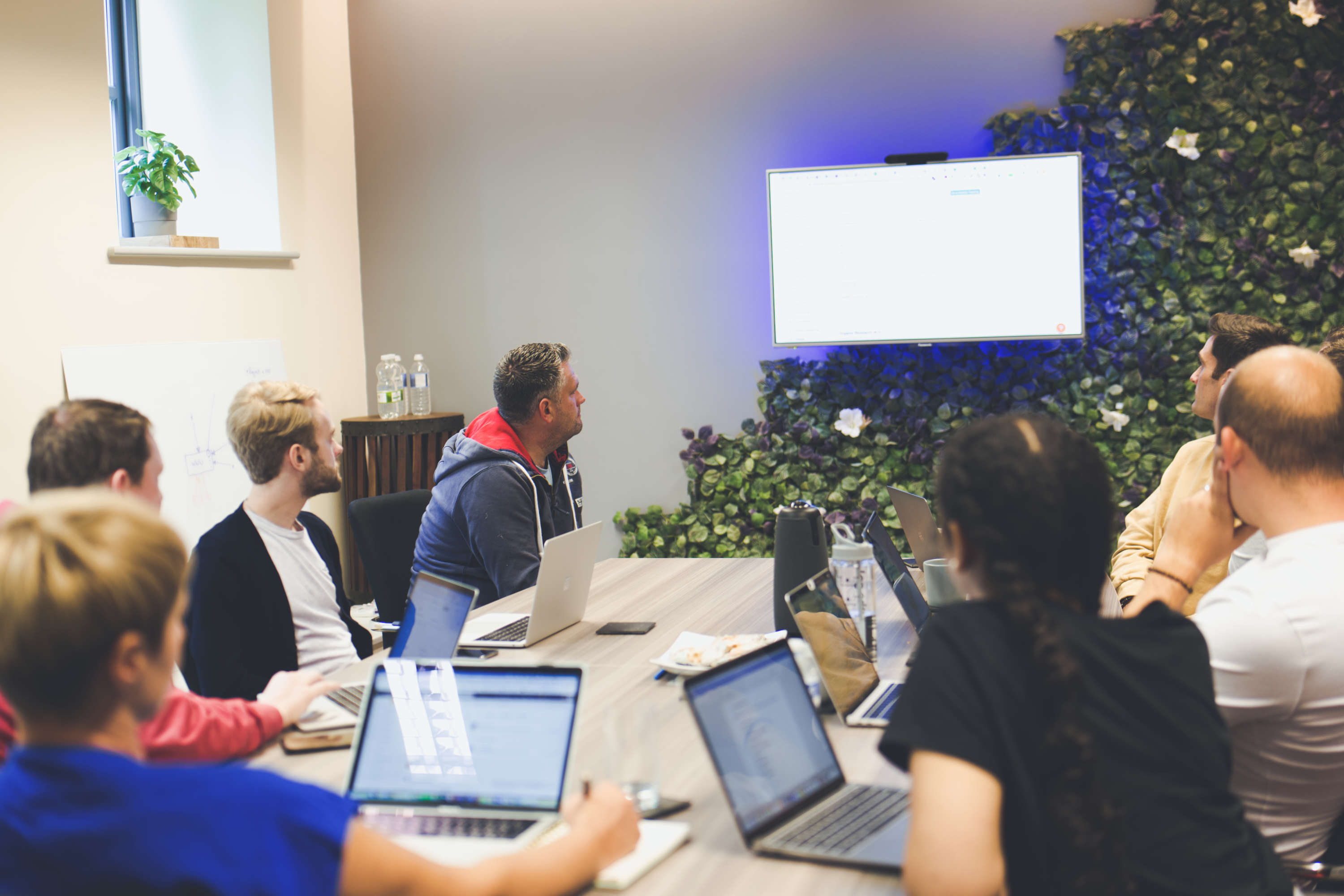 A team of digital marketers review information displayed on a large screen. All are using laptops and are focussed on the screen on the wall.