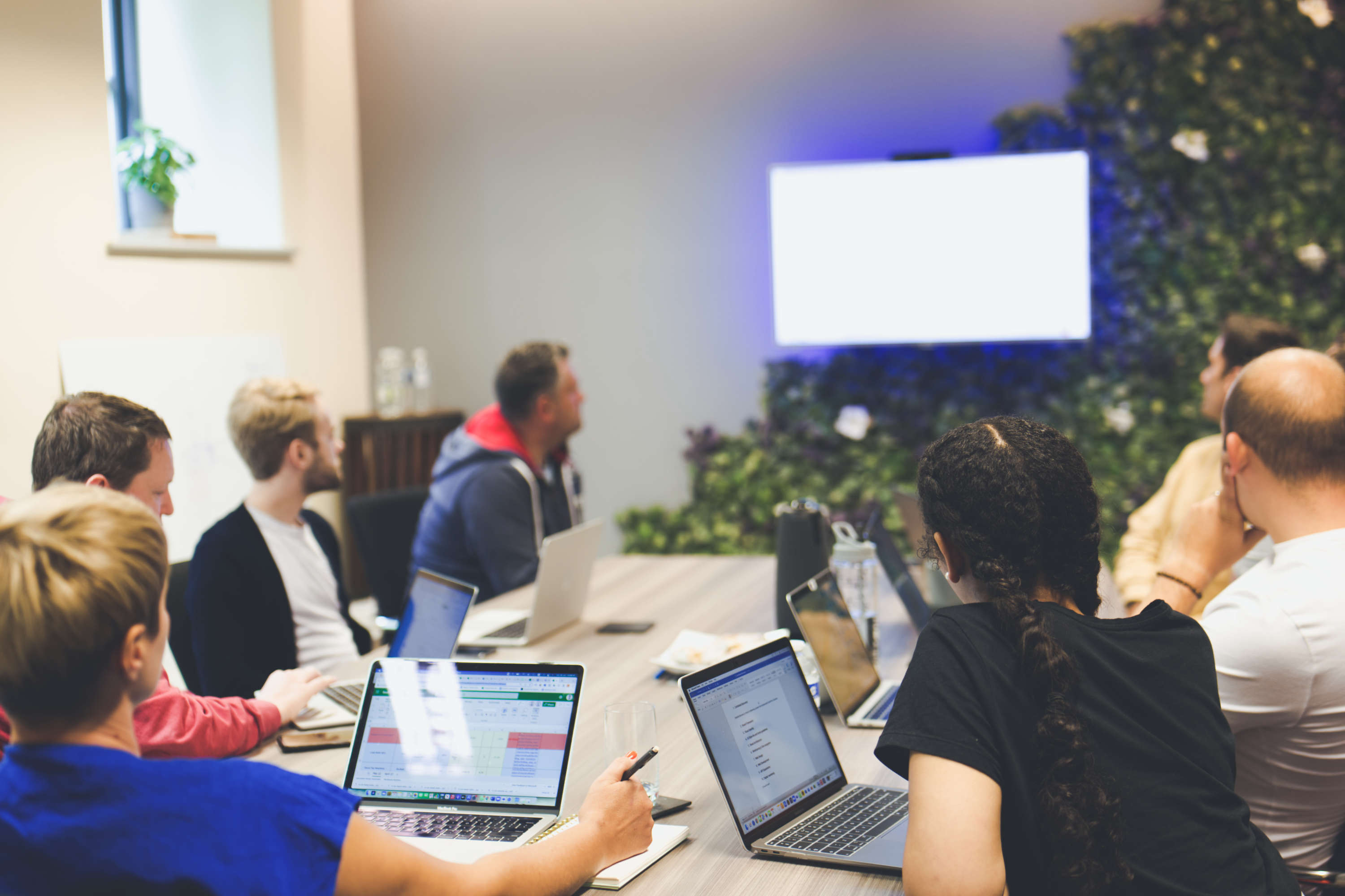 Digital marketing team members share information on a large monitor during a meeting. All are sat around a large desk and are focused on the wall mounted monitor.