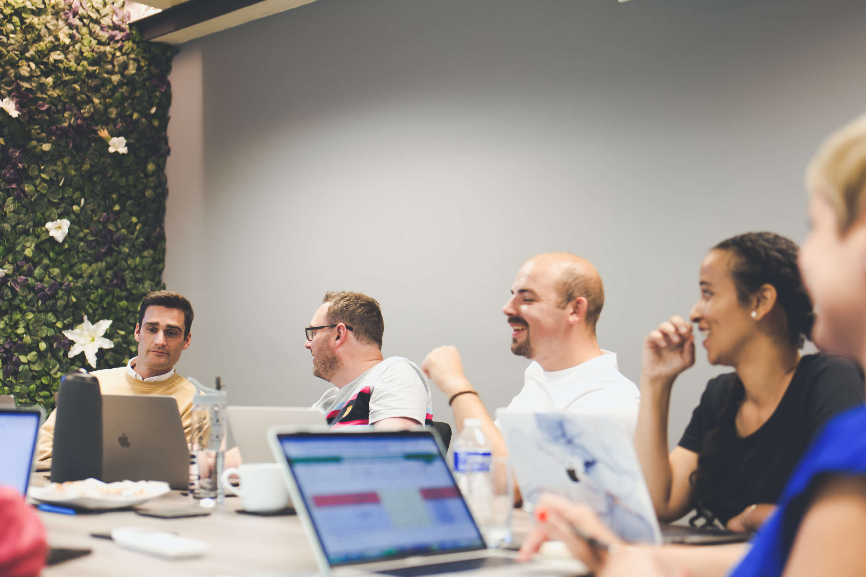 Five members of the digital marketing team conduct a team meeting around a large meeting table. All are smiling and discussing with one another.