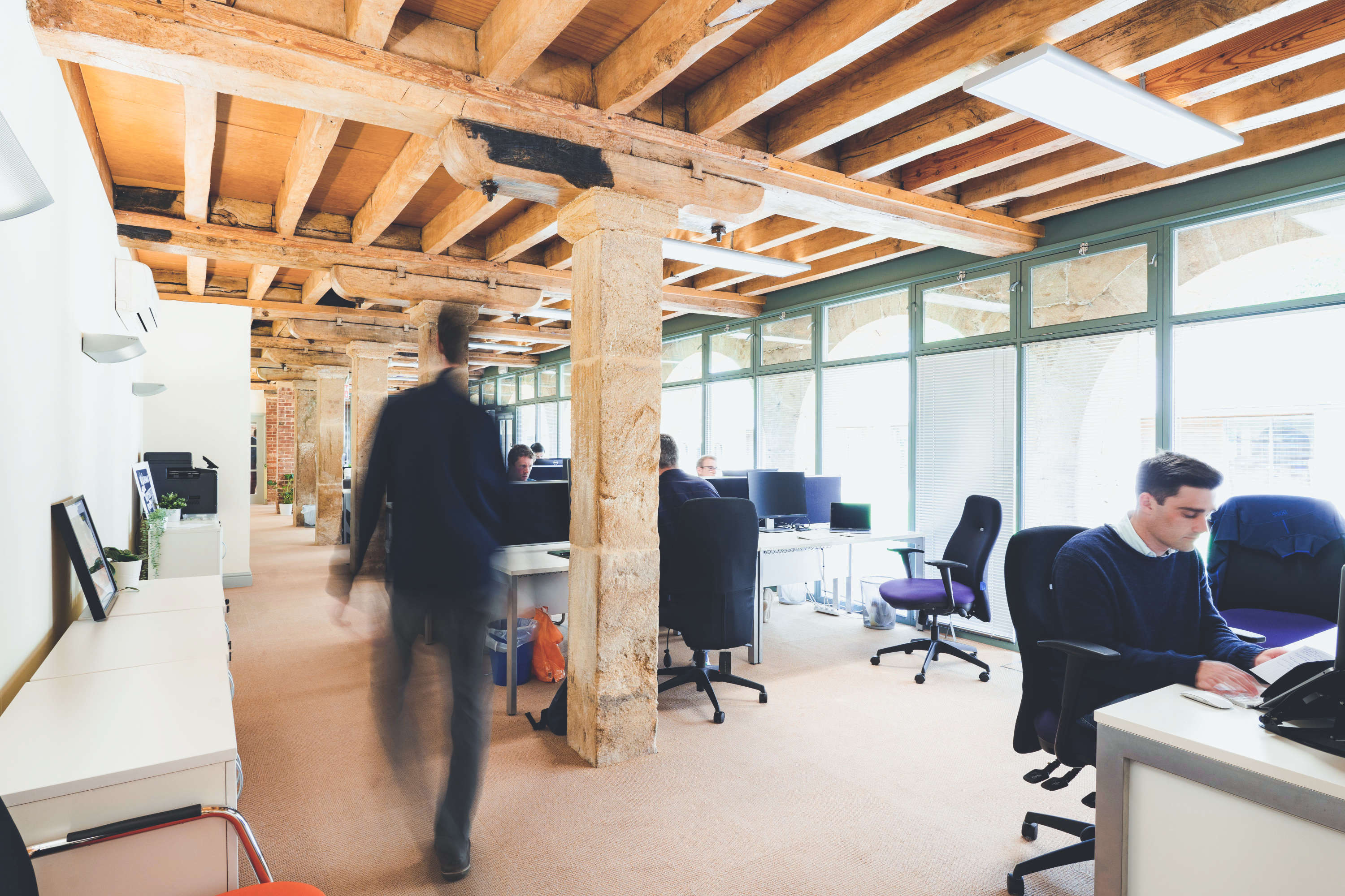 Wide-angle image of rustic modern interior space with morphsites team members working at desks. A colleague walks past the camera against a background of a large glass wall.