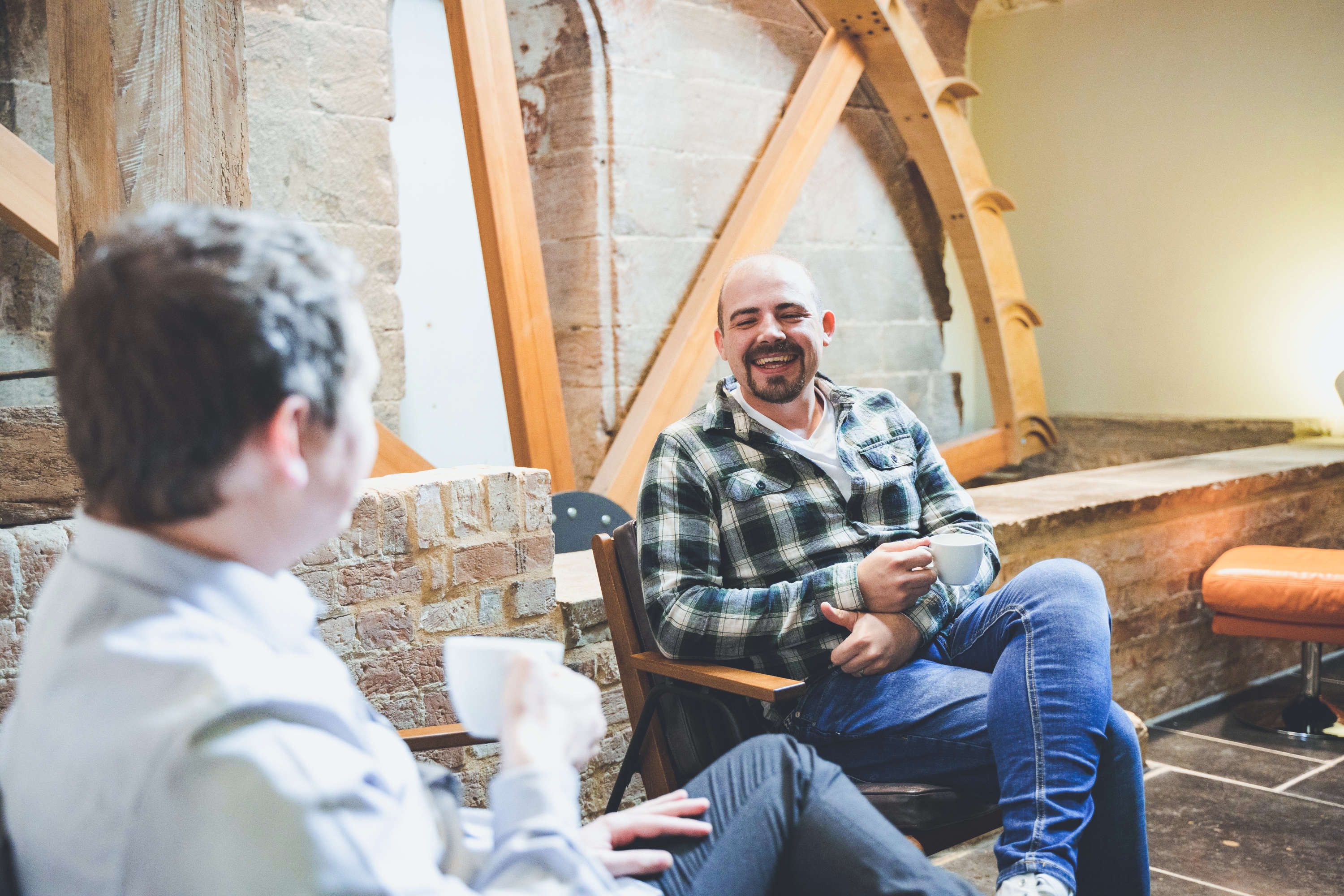 Two colleagues sit on comfy chairs in front of a rustic modern waterwheel in a social room with a relaxed studio atmosphere. Both are smiling and drinking coffee.