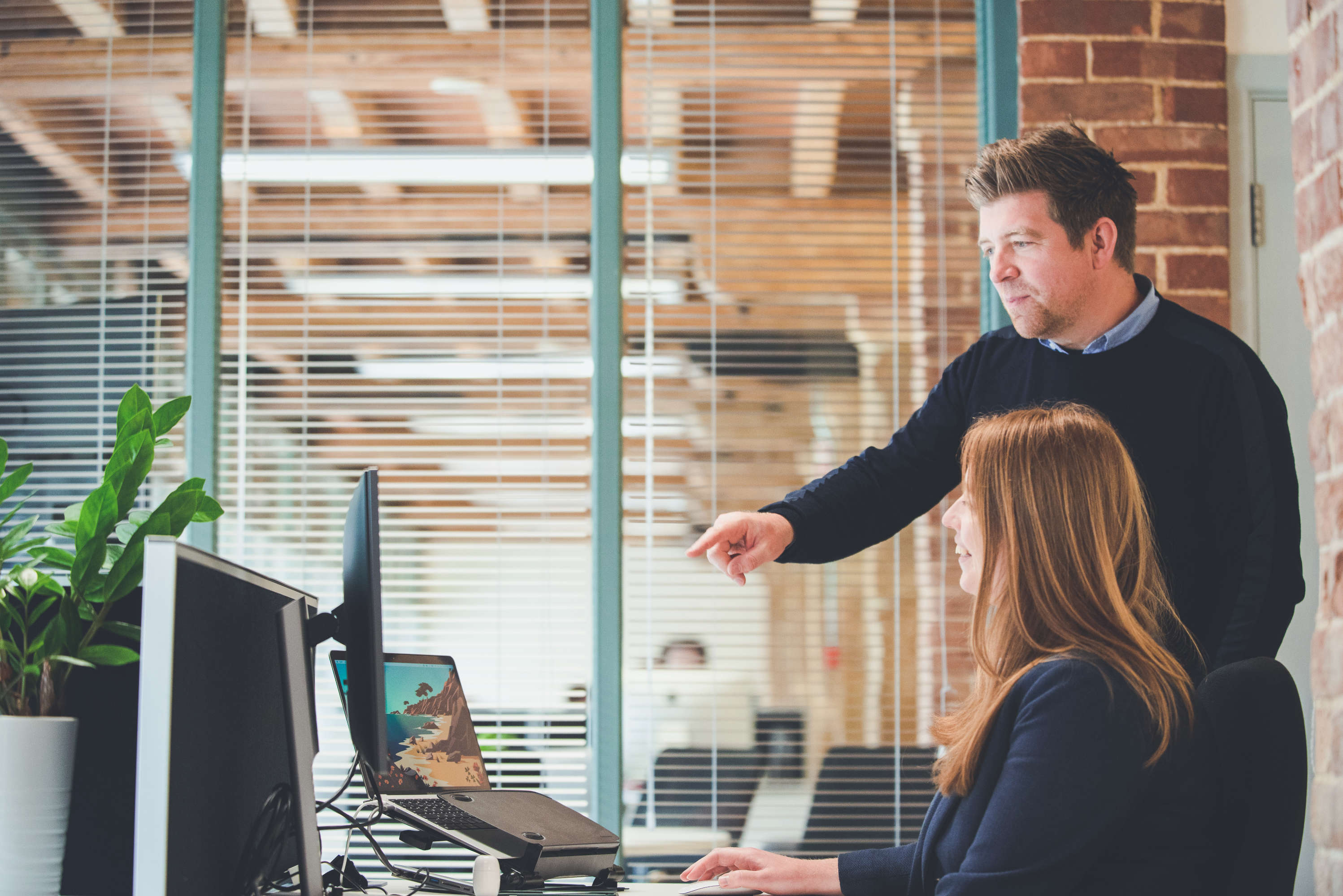 The managing director gestures to the studio managers display during a catchup. Both are smiling, and a large glass wall and brick pillar are visible in the background.