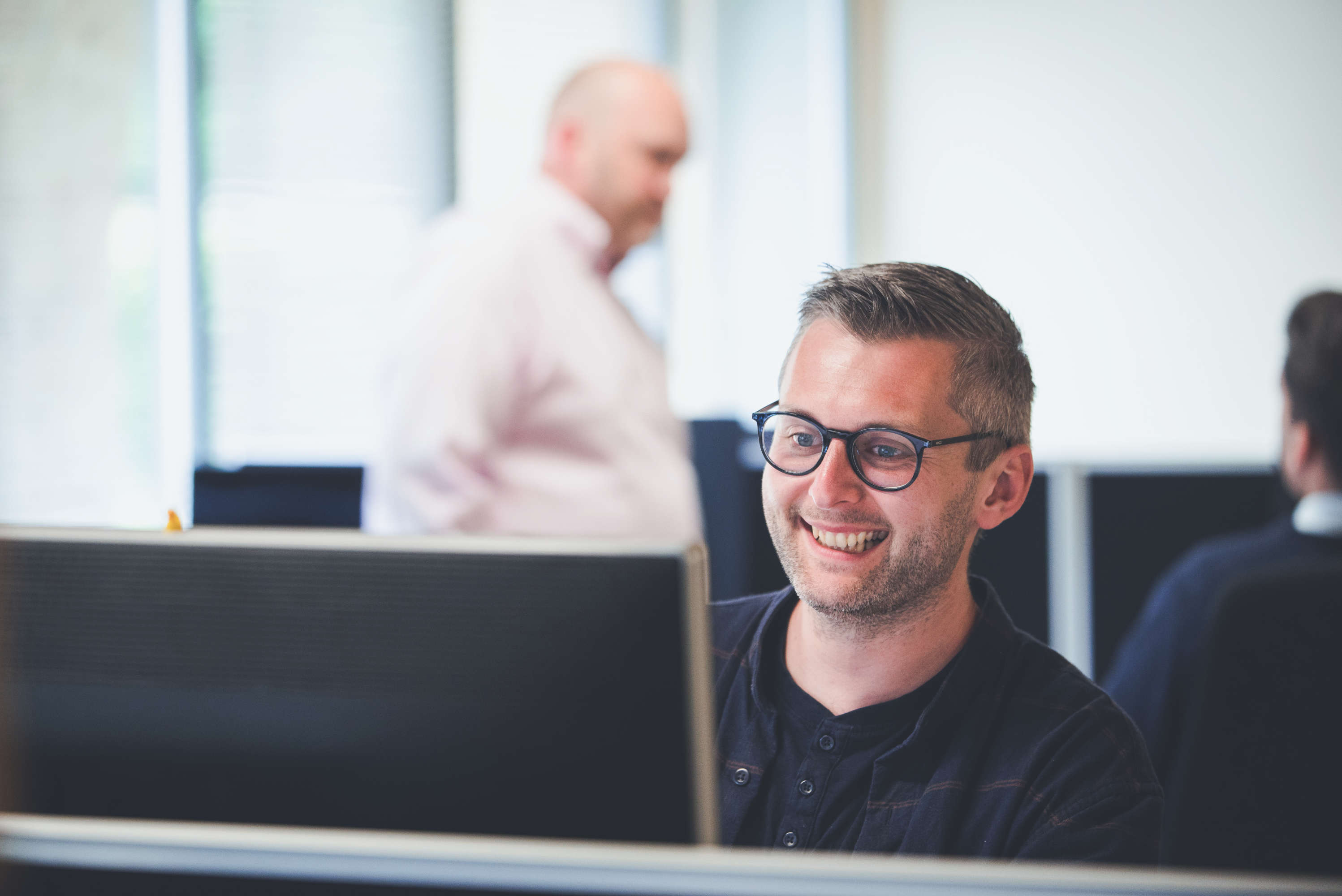 Senior Testing Technician smiles for the photographer whilst looking at his screen. Other colleagues in the background discuss a pitch.