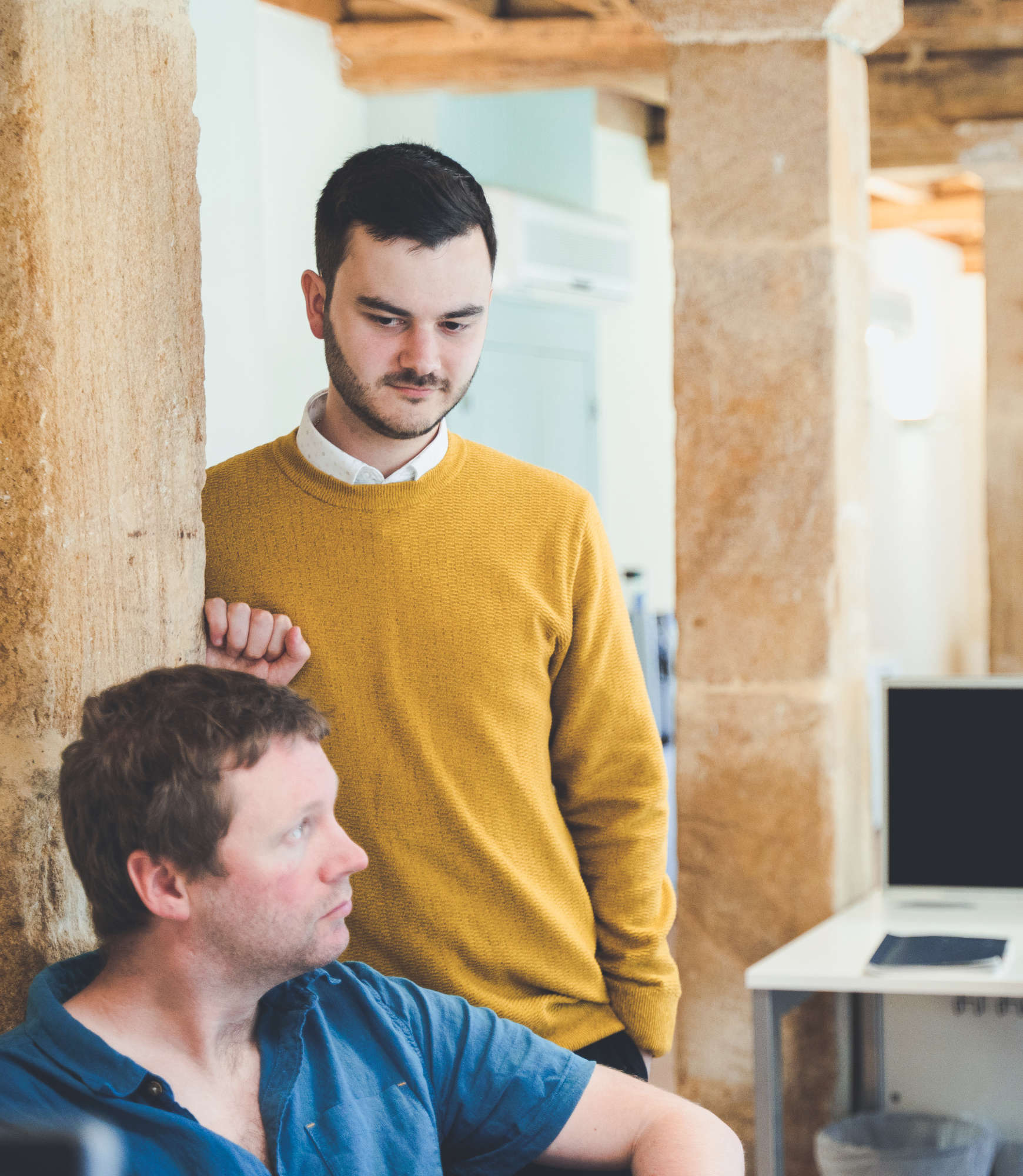 A developer and a project manager casually discuss a project, with one leaning against a stone pillar in a characterful office space. The developer looks out of frame at his colleague.