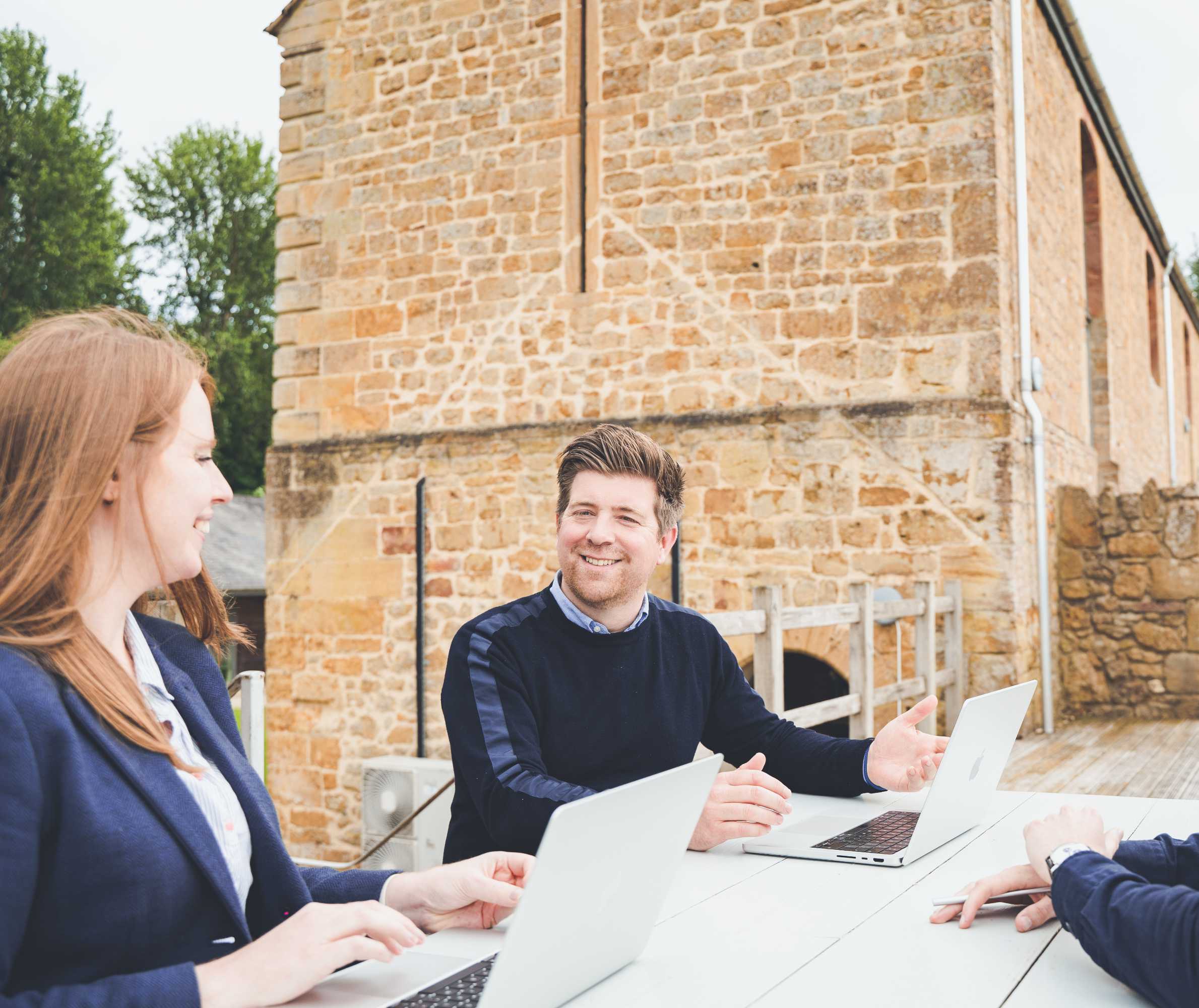 Managing director and studio manager catchup outdoors on the decking with their laptops on the table. Both are smiling and gesturing at their laptops.