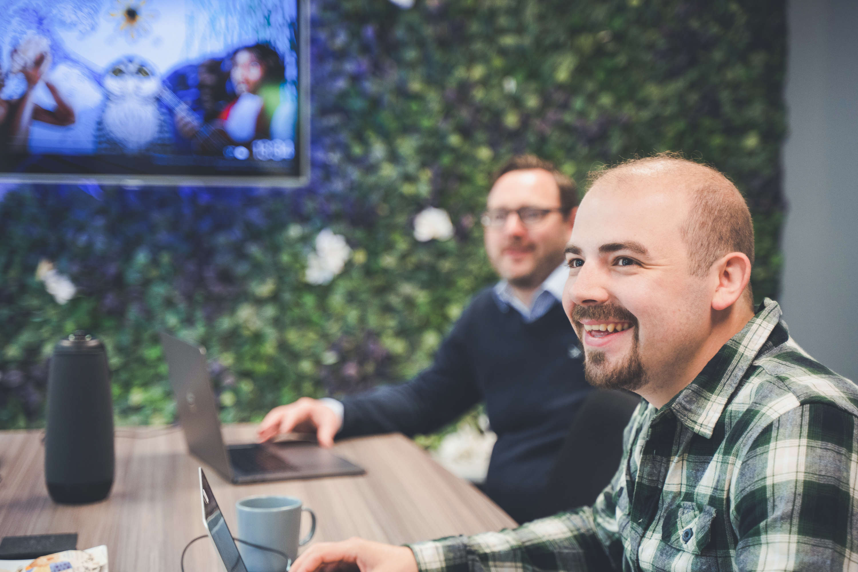 Members of the digital marketing team meet around a large table in the meeting room. Both are smiling at their colleague out of frame whilst chatting.