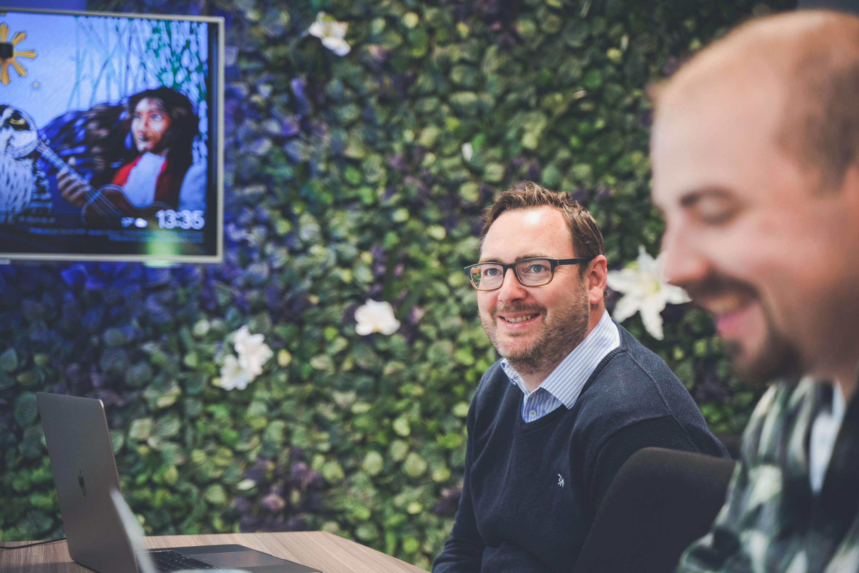 Digital marketing team members discuss around a large meeting table. Digital strategy manager looks at colleague out of frame, both are smiling.