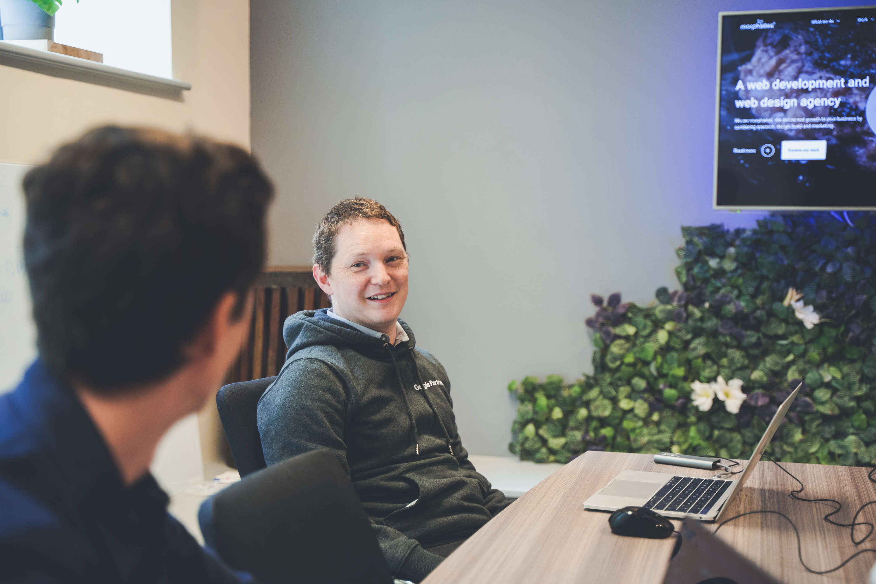 Head of digital marketing discussing work at a large desk with creative director. Both are smiling, with a large screen in the background.