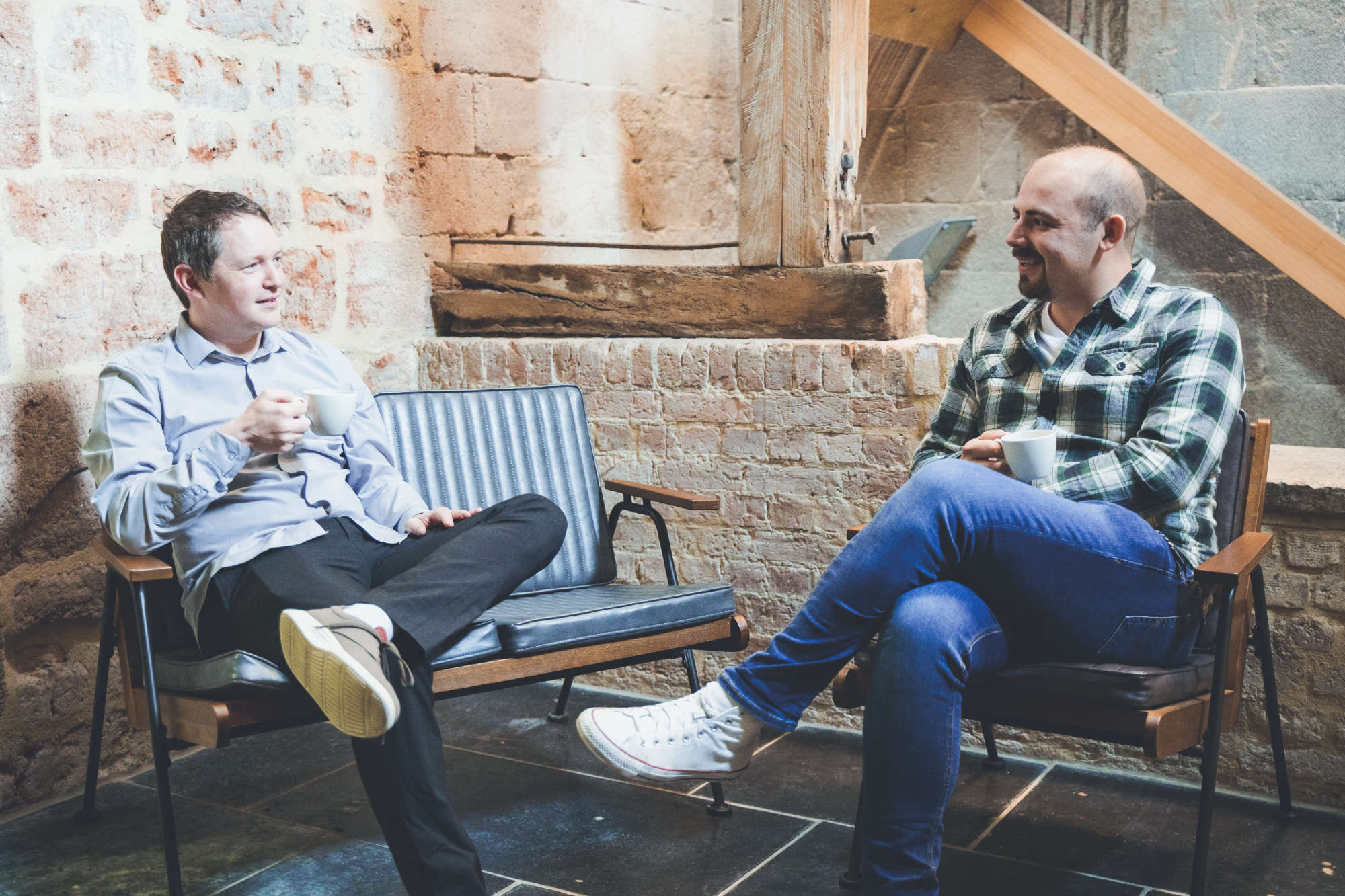 Two digital marketing team members sitting on modern retro sofas catch up over coffee in a rustic feature room.