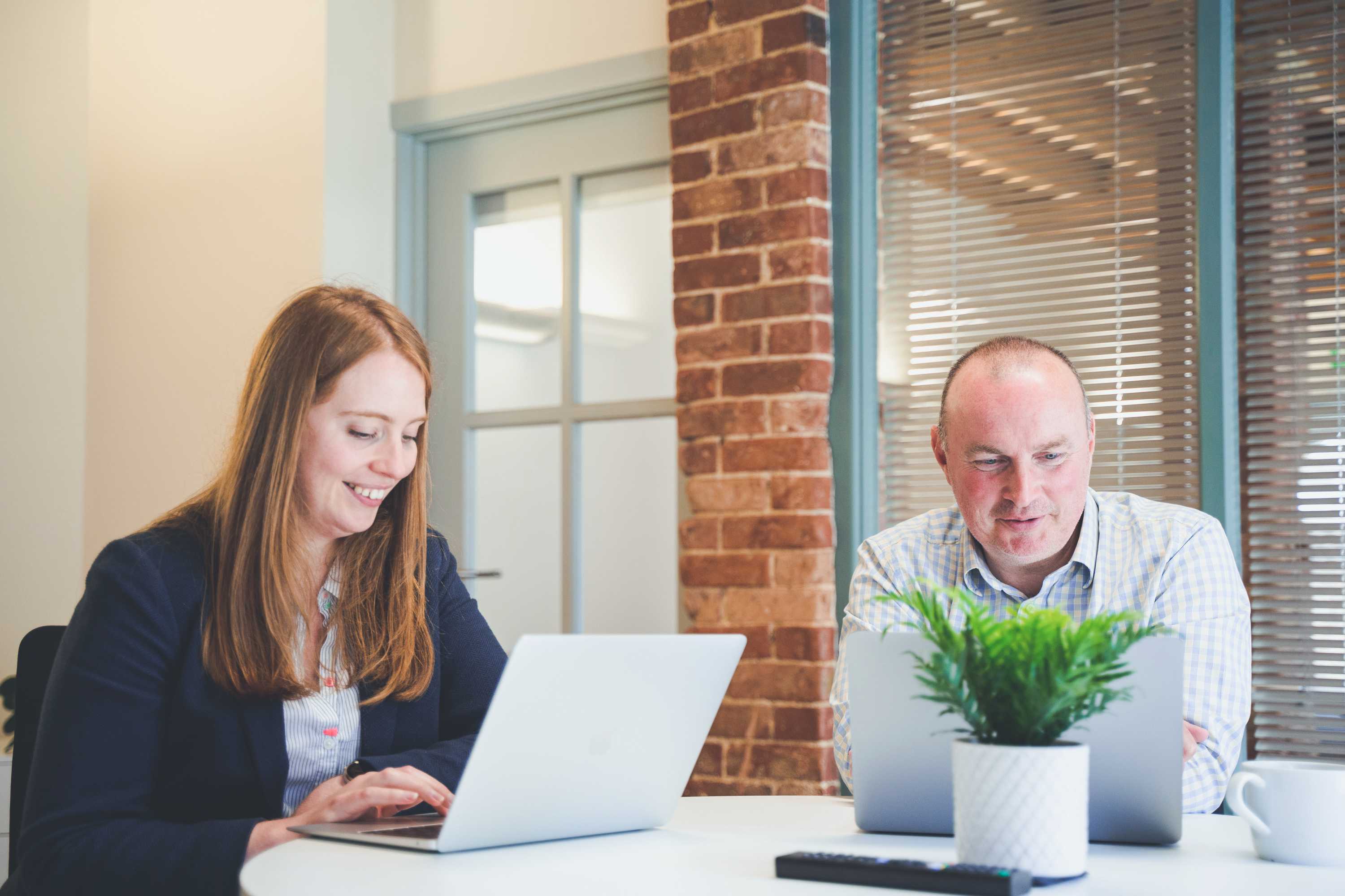 A studio manager and commercial manager catchup over coffee whilst using their laptops in a brightly lit office space.