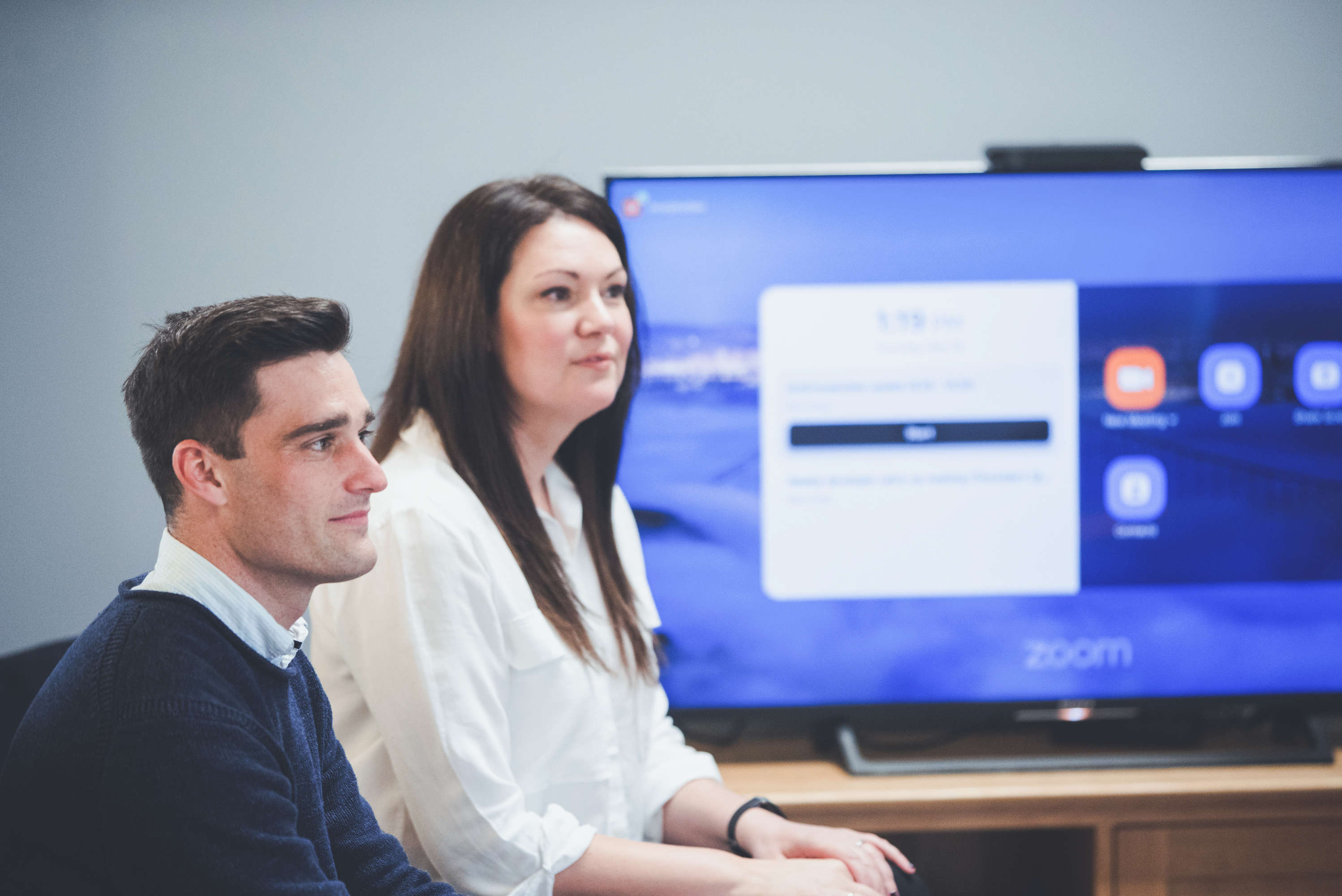 A project manager and client manager discuss with a colleague before a video conference call. A large screen behind them is set up for the video call, both are focusing on their colleague who is out of frame.