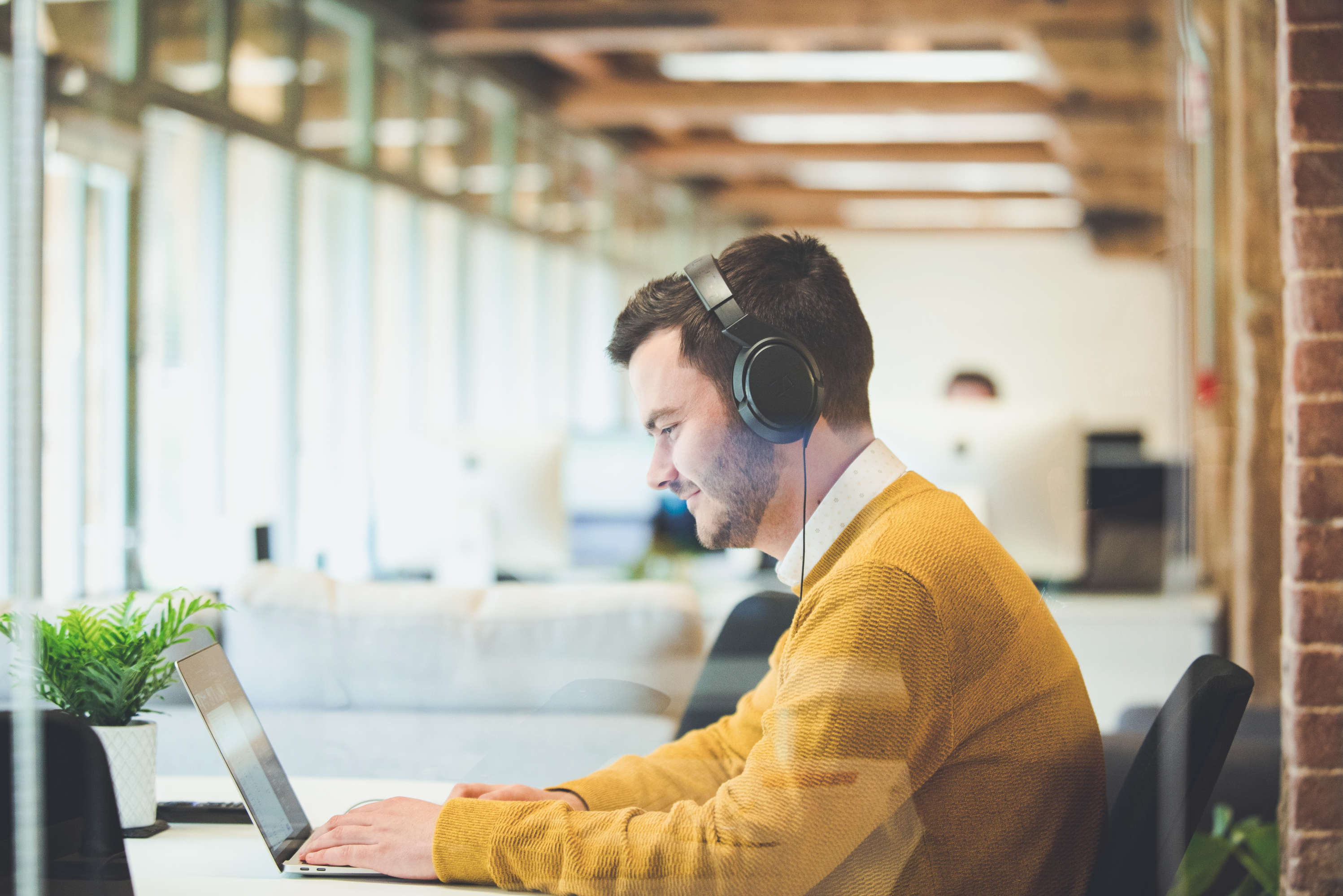 A project manager responds to client requests on his laptop, in a bright, open-plan office space. Other team members are working in the background.