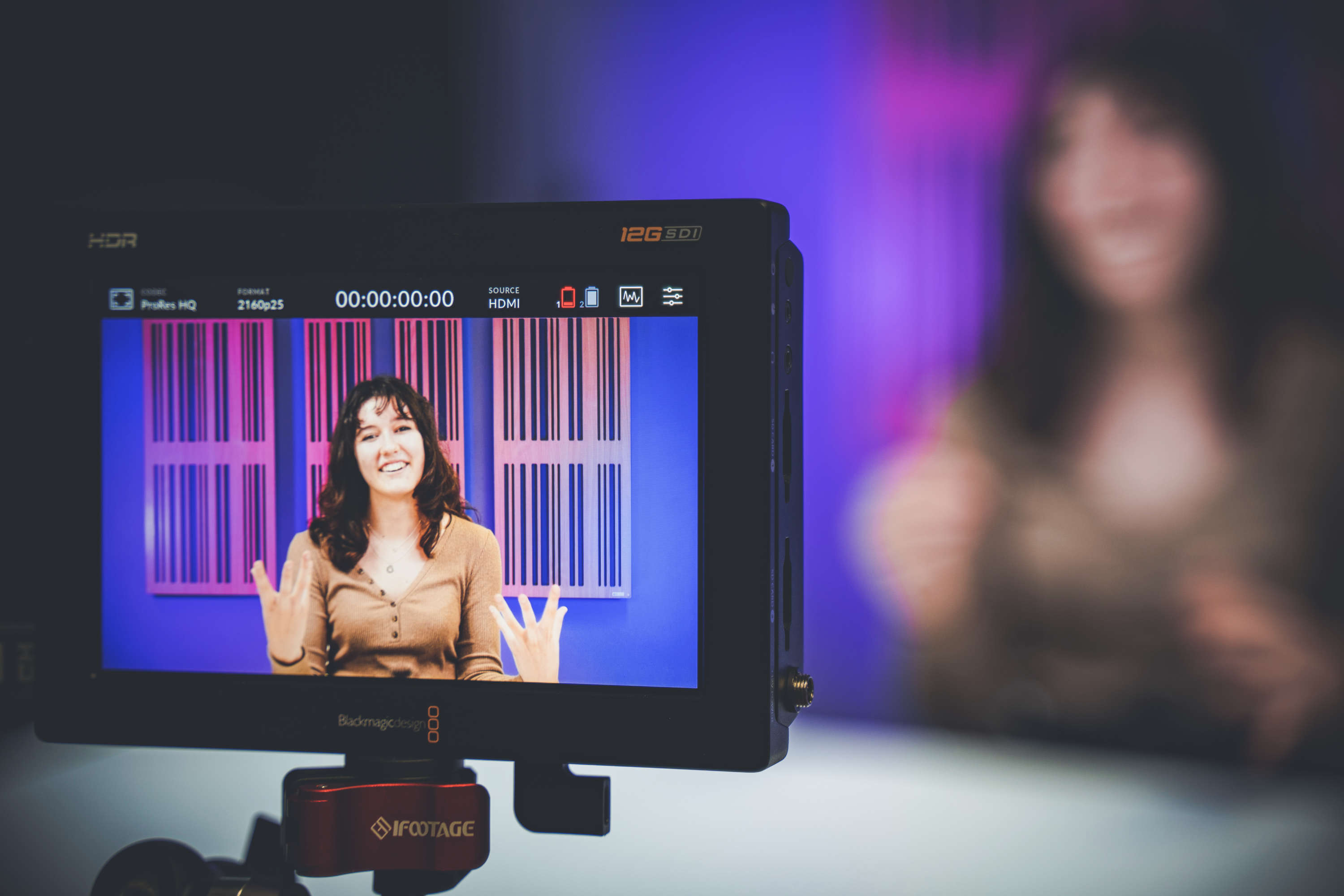 A video presenter gestures to the camera, viewed through a camera connected monitor. The presenter is also visible in the background, but out of focus.
