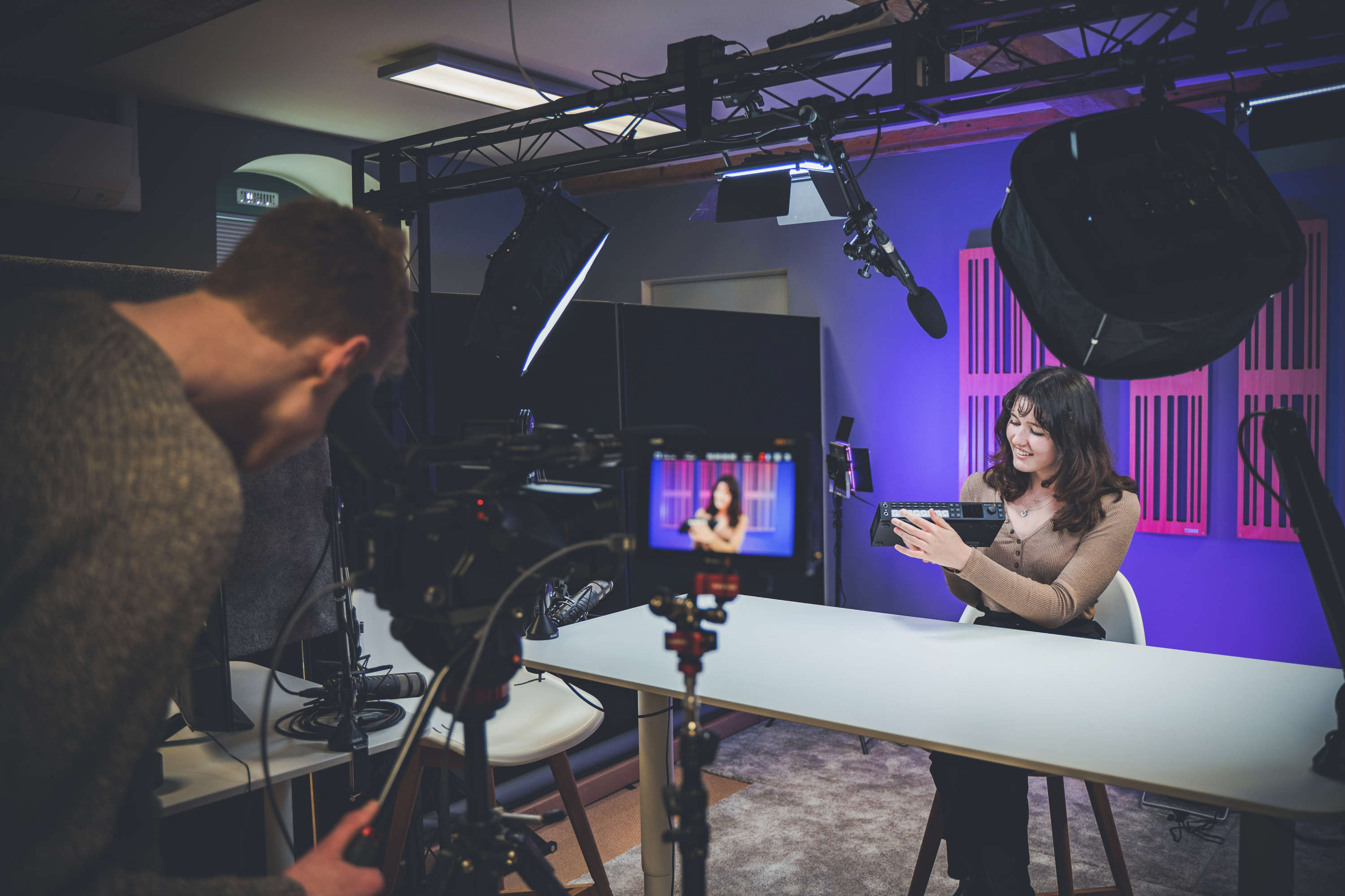 A presenter shows off a product whilst recording a product review. The presenter points to a feature of the product whilst a camera operator checks the shot framing.
