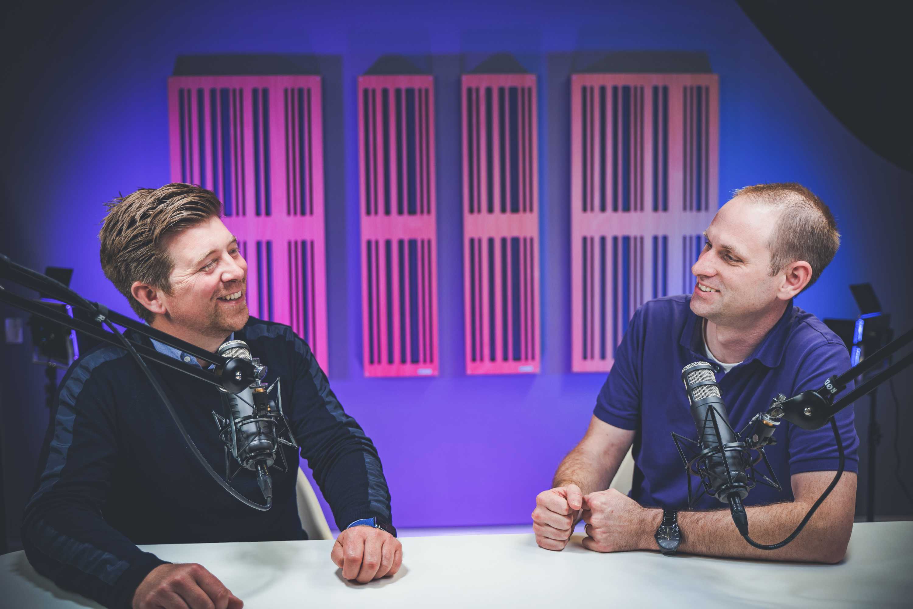 Two presenters smile at each other whilst filming a new video. Both are leaning back, and are surrounded by studio equipment.