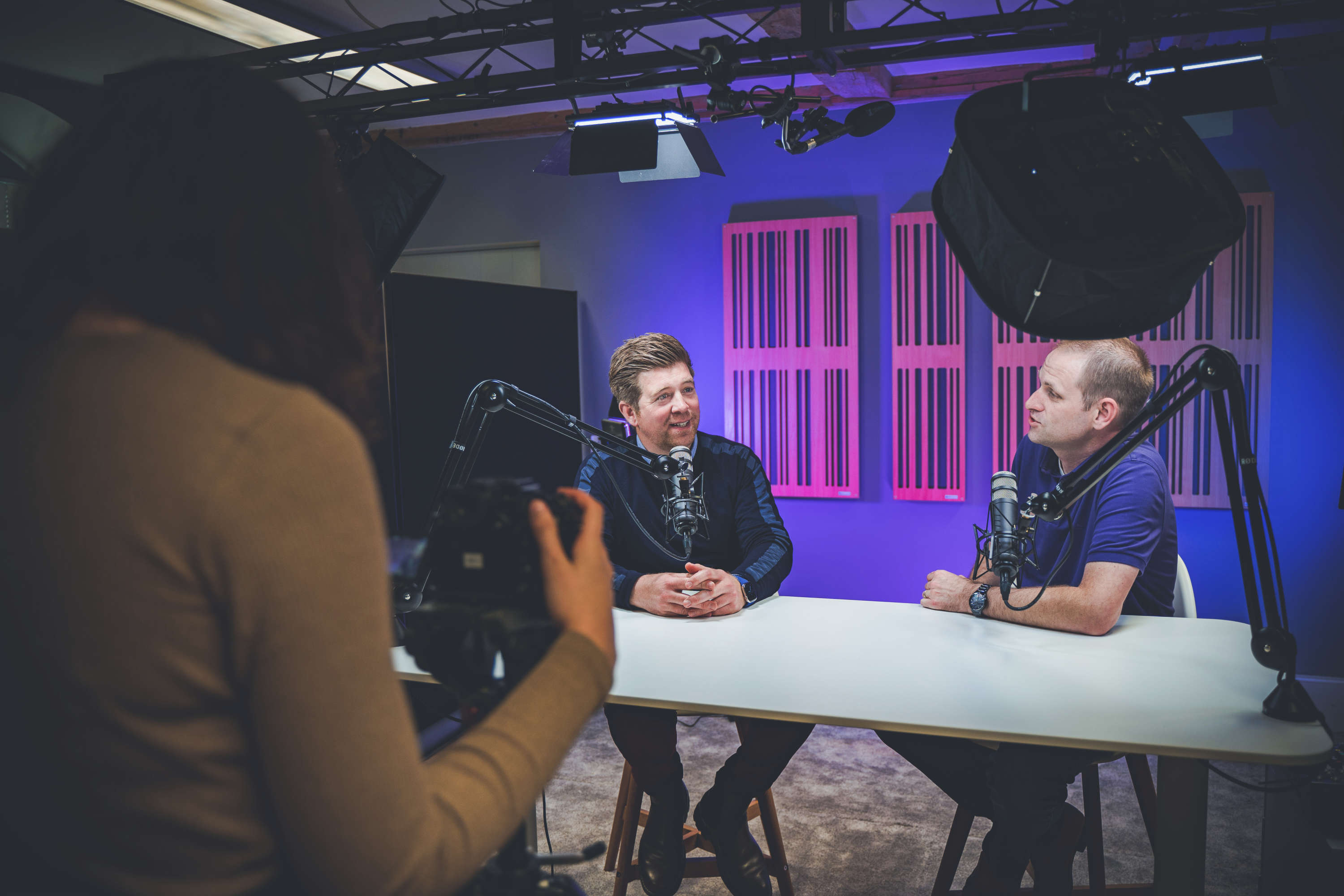 Two podcast presenters record an informal discussion. A camera operator takes images using a mounted camera.