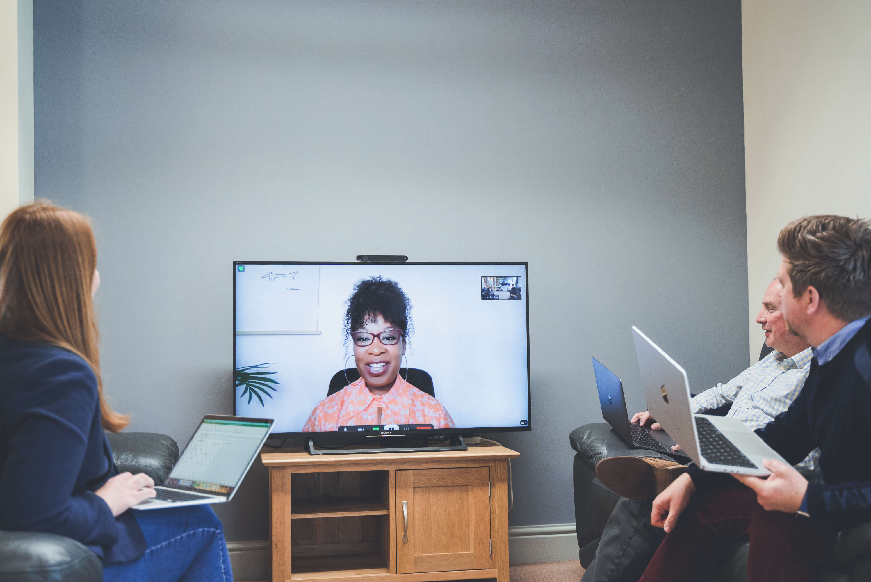 Marketing manager conducts a video call meeting with senior management team, appearing on a large monitor.