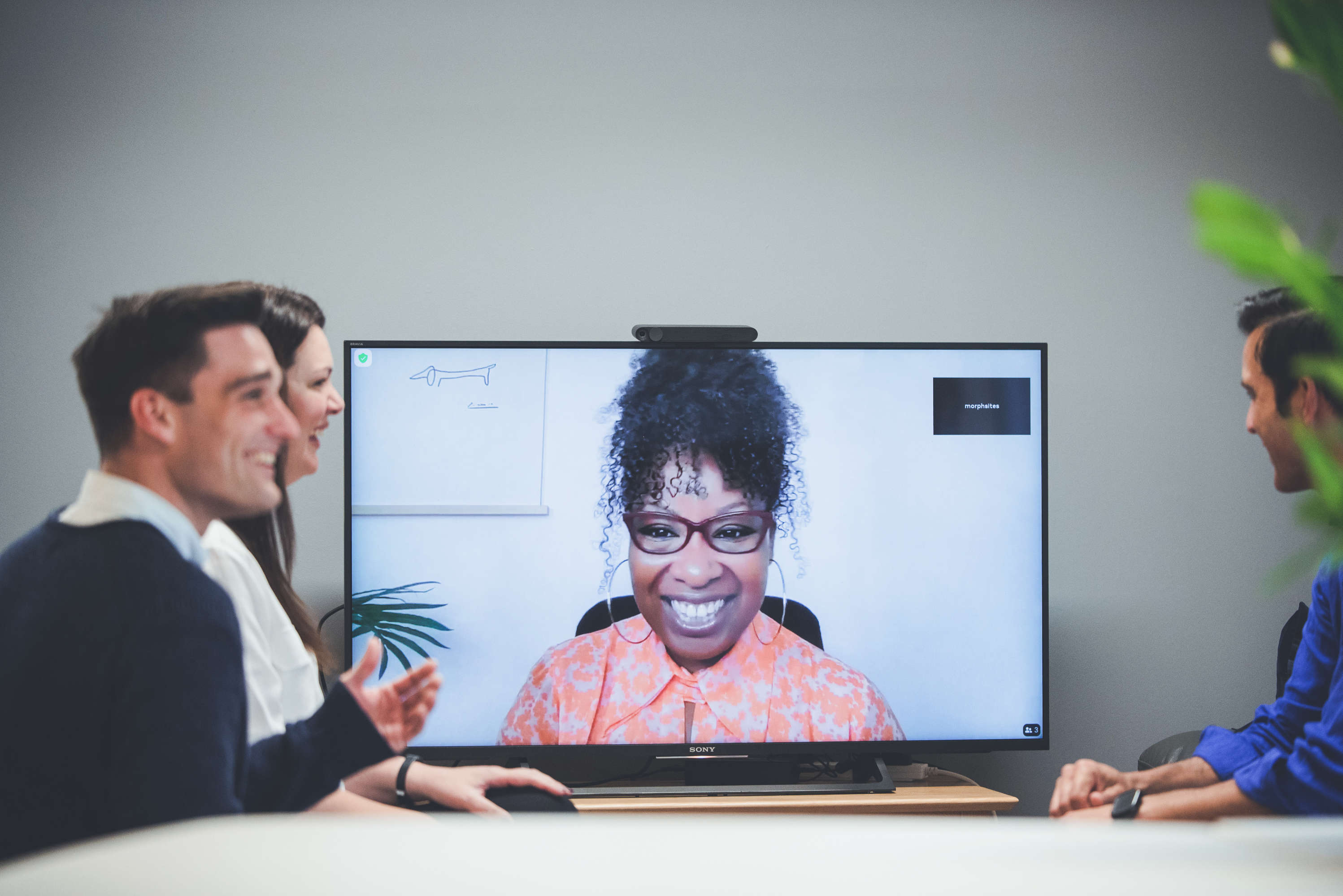 Project and client management team members catch up with a colleague via video call who appears on a large screen.