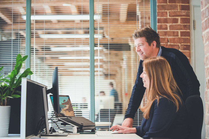 Managing director and studio manager at a desk both look at a screen, both are smiling.