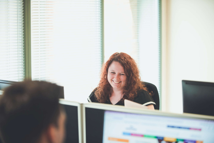 Accountant looking at their screen smiles whilst chatting with support team colleagues.