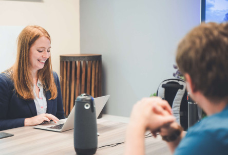 A project manager and developer sat at a large table chat before a meeting, both are smiling.