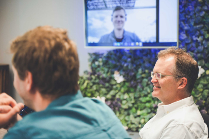 Two senior developers relax and smile during a team meeting. A screen in the background shows a colleague via video call.
