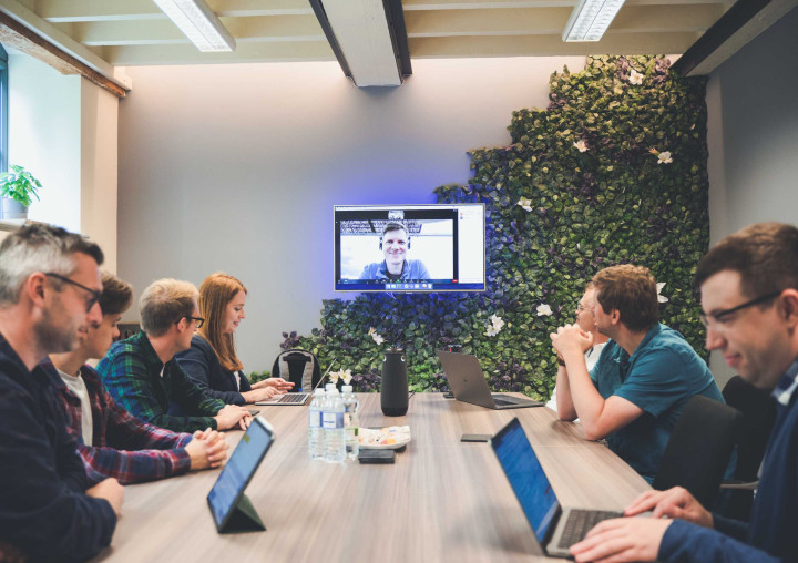 A team of seven web developers and a project manager conduct a video call meeting with a colleague around a large desk. Some attendees are looking at laptops, others up at the screen.