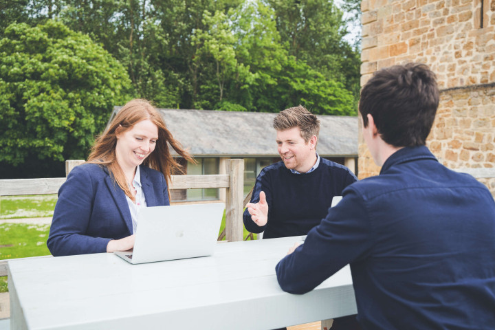 Managing director, studio manager and creative director chat outside around a bench table. Office buildings are surrounded by trees in the background.
