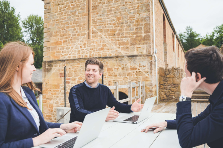 The managing director, studio manager and creative director catchup outside. All are sitting around a bench table on a sunny deck, smiling.