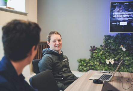 Head of digital marketing discussing work at a large desk with creative director. Both are smiling, with a large screen in the background.