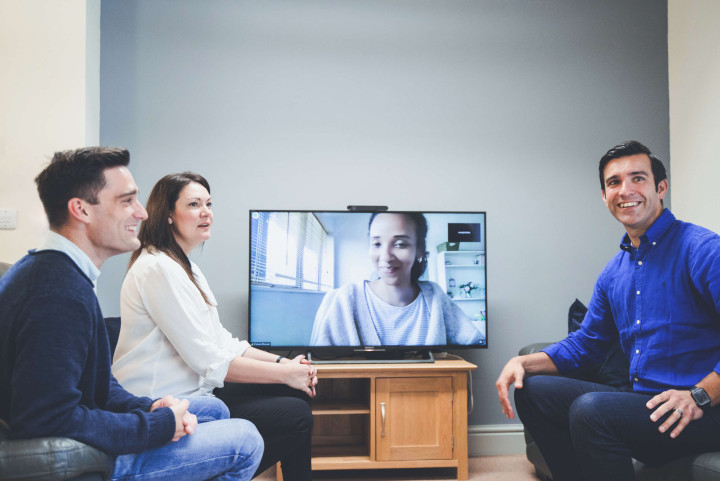 Whilst relaxing on sofas, the project management team and client manager look at the camera during a video call with a remote working colleague. All are smiling or laughing.